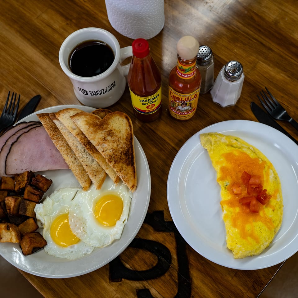 Breakfast plates with eggs, ham, toast, potatoes, and an omelet sit on a wooden table with coffee and hot sauce.