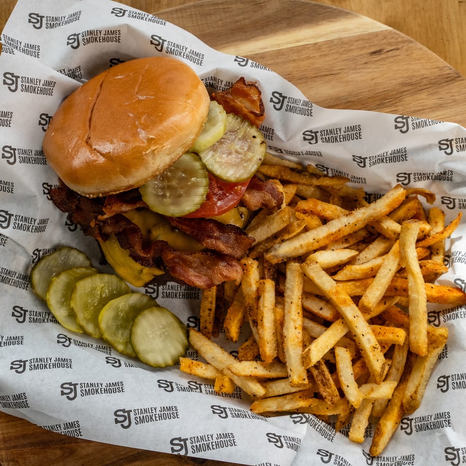 Bacon cheeseburger with pickles, tomato, and seasoned fries served on branded Stanley James Smokehouse paper.