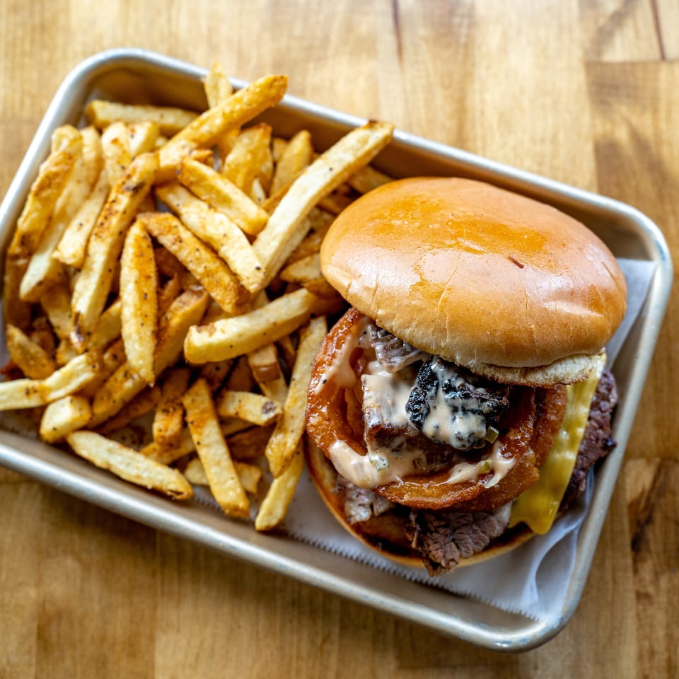 Loaded cheeseburger with bacon, sauce, and pickles served alongside seasoned French fries on a metal tray.