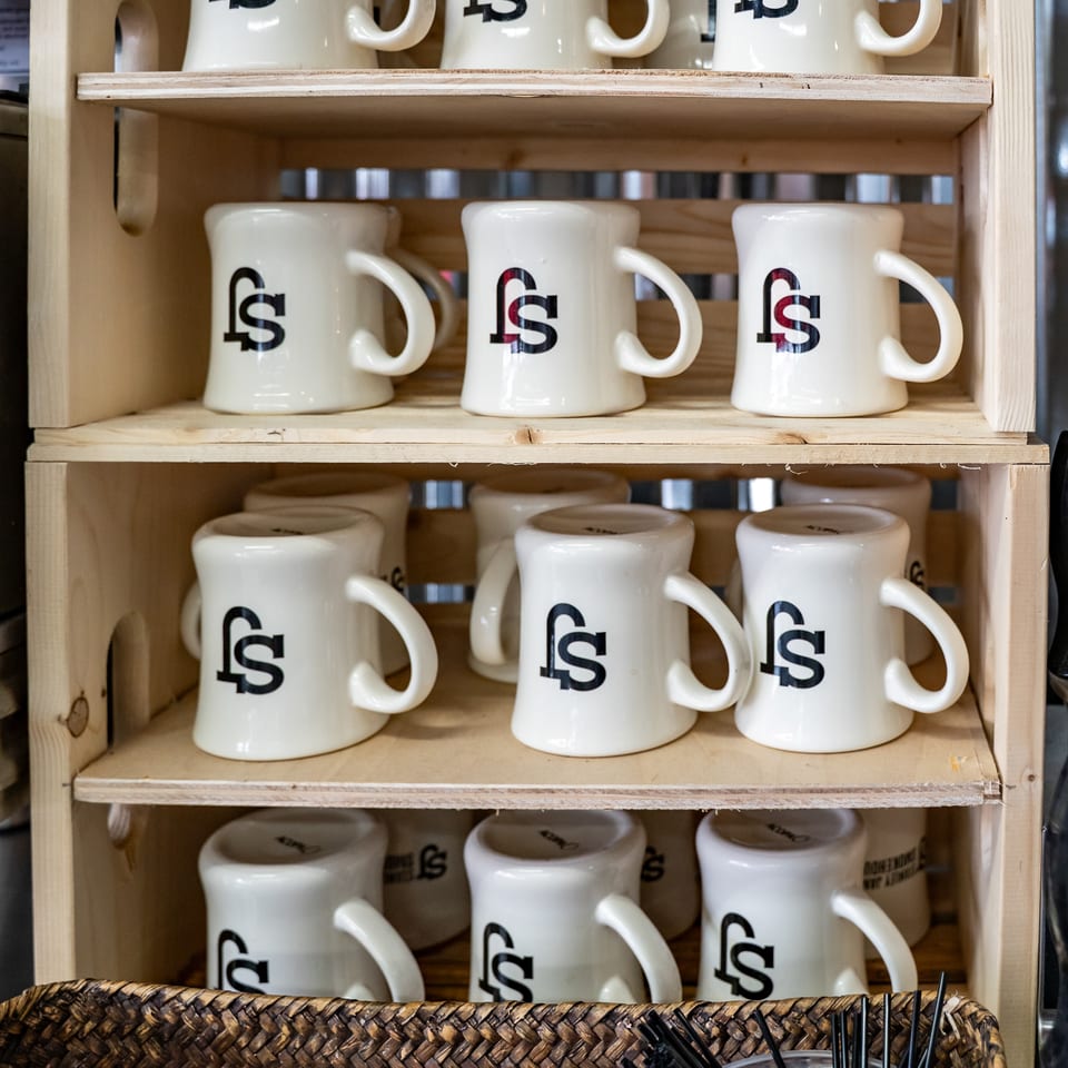 White branded ceramic mugs arranged on wooden shelves above woven baskets holding takeaway lids and black straws.