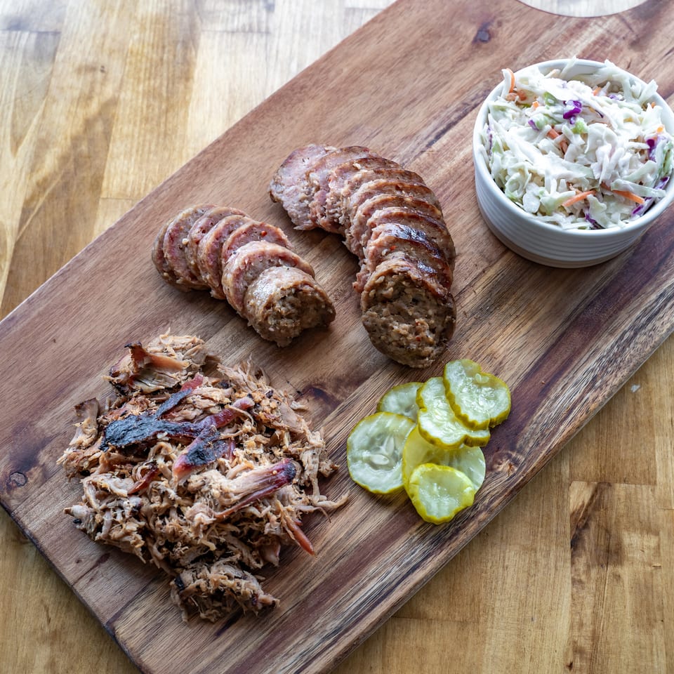 Sliced sausages, pulled pork, coleslaw, and pickles arranged on a wooden serving board.
