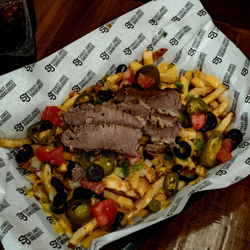 Loaded fries topped with sliced smoked beef, melted cheese, jalapeños, olives, tomatoes, peppers, and onions beside a soft drink.