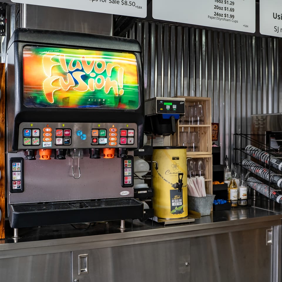 Restaurant beverage station with a colorful soda fountain, ice dispenser, coffee machines, cups, and drink pricing signs.