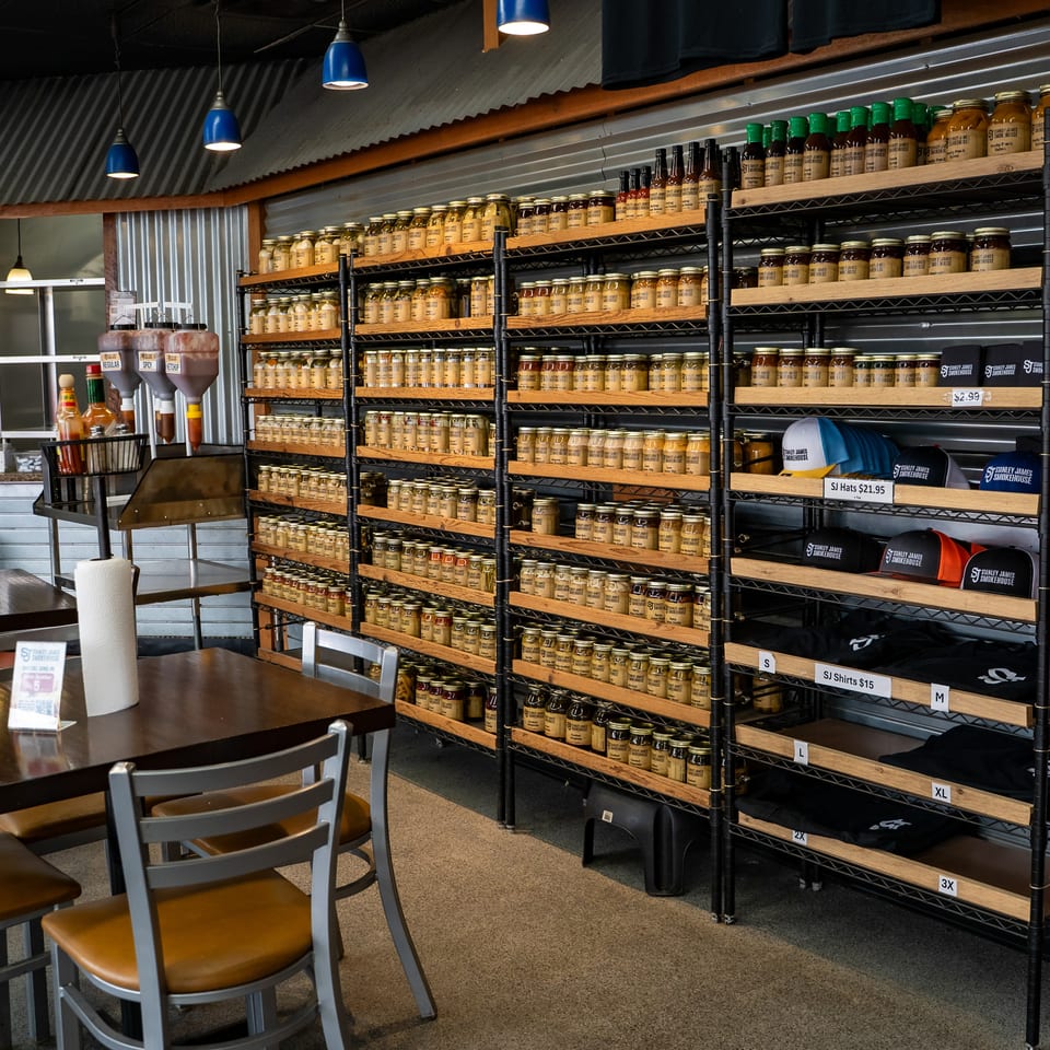Stanley James Smokehouse dining area with tables, condiment station, and shelves stocked with jars, hats, and branded clothing.