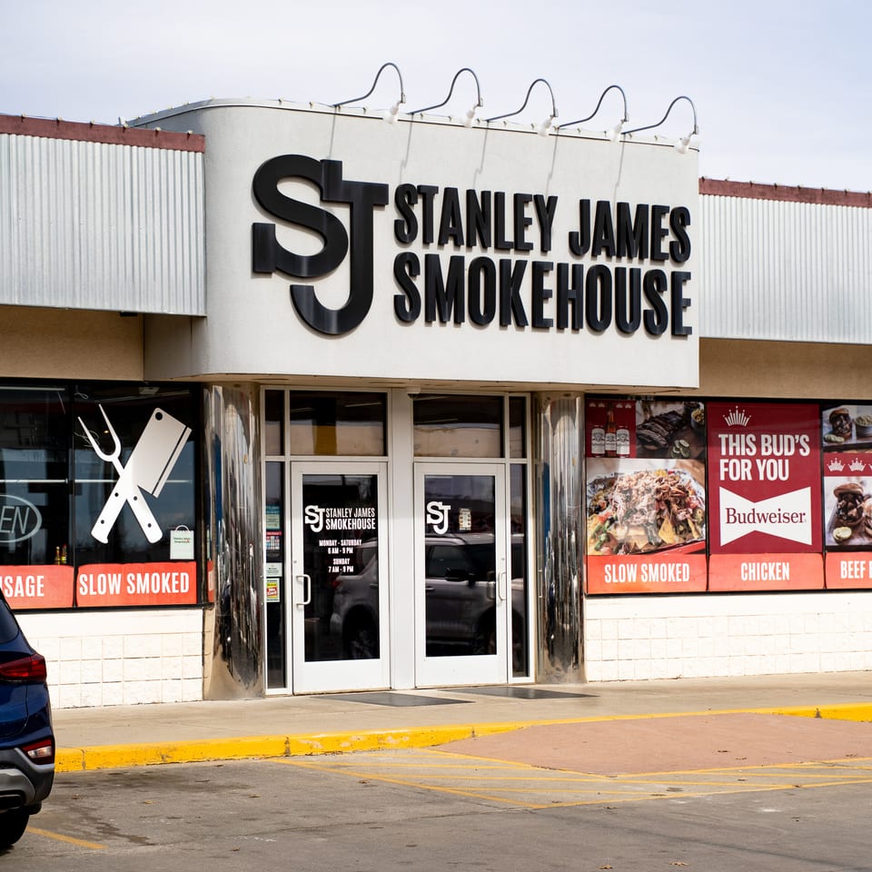 Exterior of Stanley James Smokehouse restaurant with prominent signage, food advertisements, glass entrance doors, and a parking lot.