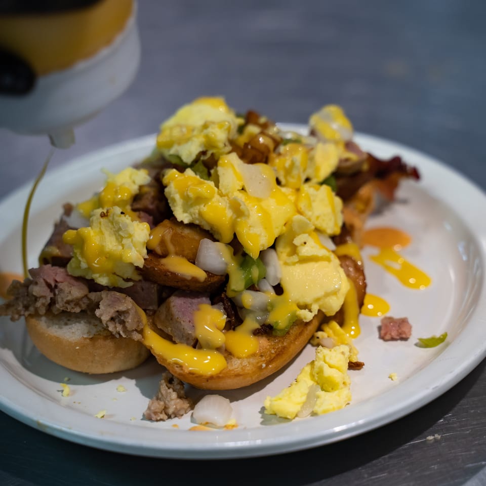 Loaded breakfast plate with sausage, scrambled eggs, potatoes, onions, and yellow cheese sauce on a white dish.
