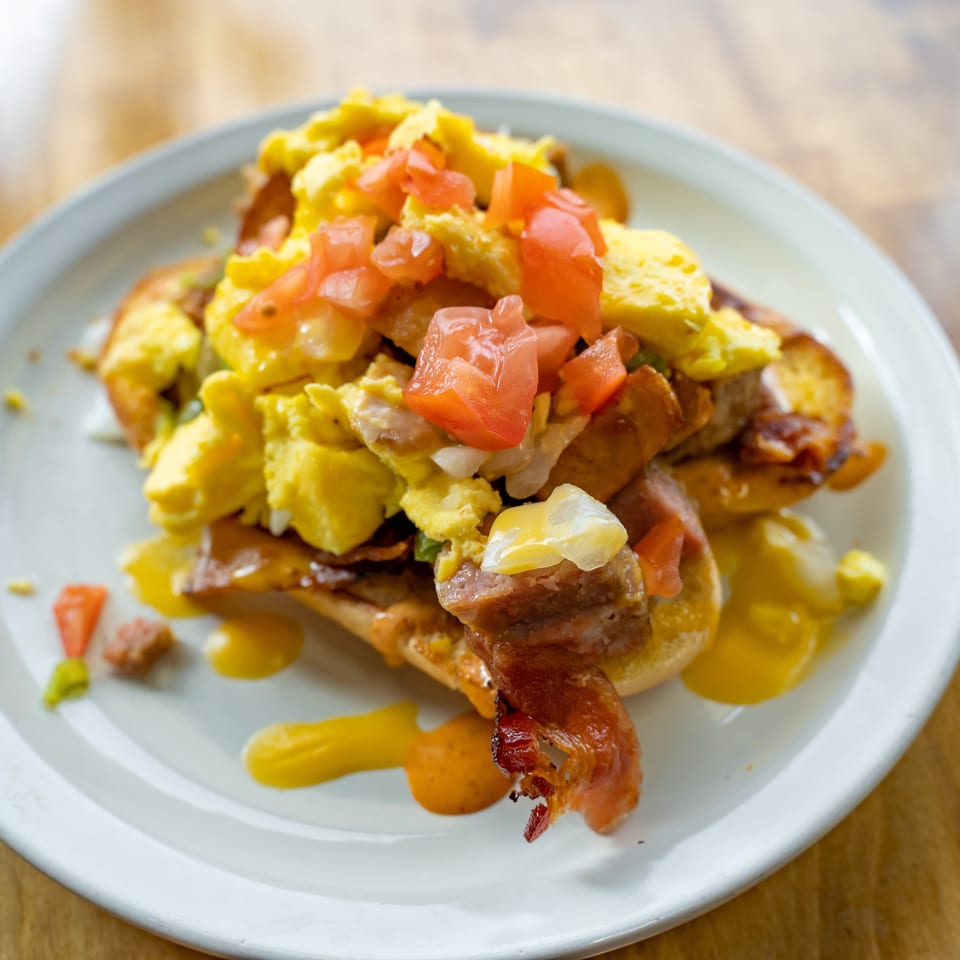 Loaded breakfast plate with scrambled eggs, bacon, sausage, diced tomatoes, avocado, and sauces on a white plate.
